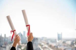 Şehir manzarası ve açık gökyüzü önünde, başarıyla tamamlanan hukuki eğitimi simgeleyen iki kırmızı kurdeleli diploma rulosunu havaya kaldıran eller.