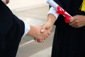 Doçentlik başvurusu sonrası tokalaşan iki akademisyen – İtiraz, jüri değerlendirmesi ve yeniden başvuru sürecini simgeleyen diploma ve cübbe detaylı görsel.