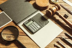 Ahşap bir masa üzerinde, hukuki muhasebeyi ve finansal davaları temsil eden bir tokmak, hesap makinesi, büyüteç ve not defteri.