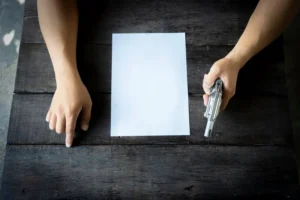Karanlık ahşap bir masa üzerinde, boş bir kağıda doğru işaret eden bir el ve yanında duran bir tabancayla ceza hukuku davalarındaki ciddi durumu temsil eden görsel.